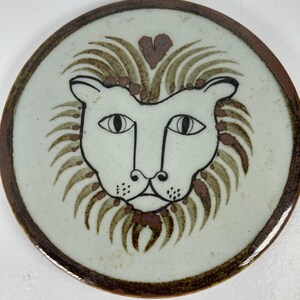 May include: A white ceramic plate with a brown rim features a hand-painted lion face with a heart above its head. The lion's mane is depicted in brown and green hues.