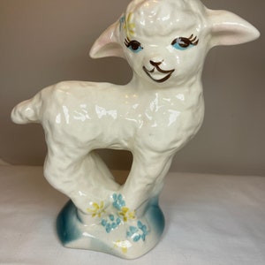 May include: A white ceramic lamb figurine with blue and yellow flowers painted on the base. The lamb has a smiling face with brown eyes and eyelashes.