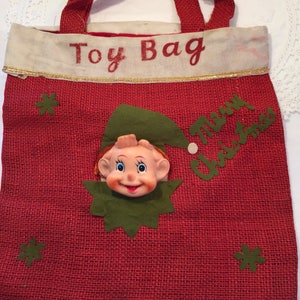 May include: Red burlap Christmas toy bag with a smiling elf face and the words "Toy Bag" and "Merry Christmas". The bag has red handles and green snowflake accents.