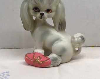 May include: A white ceramic poodle figurine with a pink heart-shaped pillow with a gold bow. The poodle has a sad expression and large brown eyes.