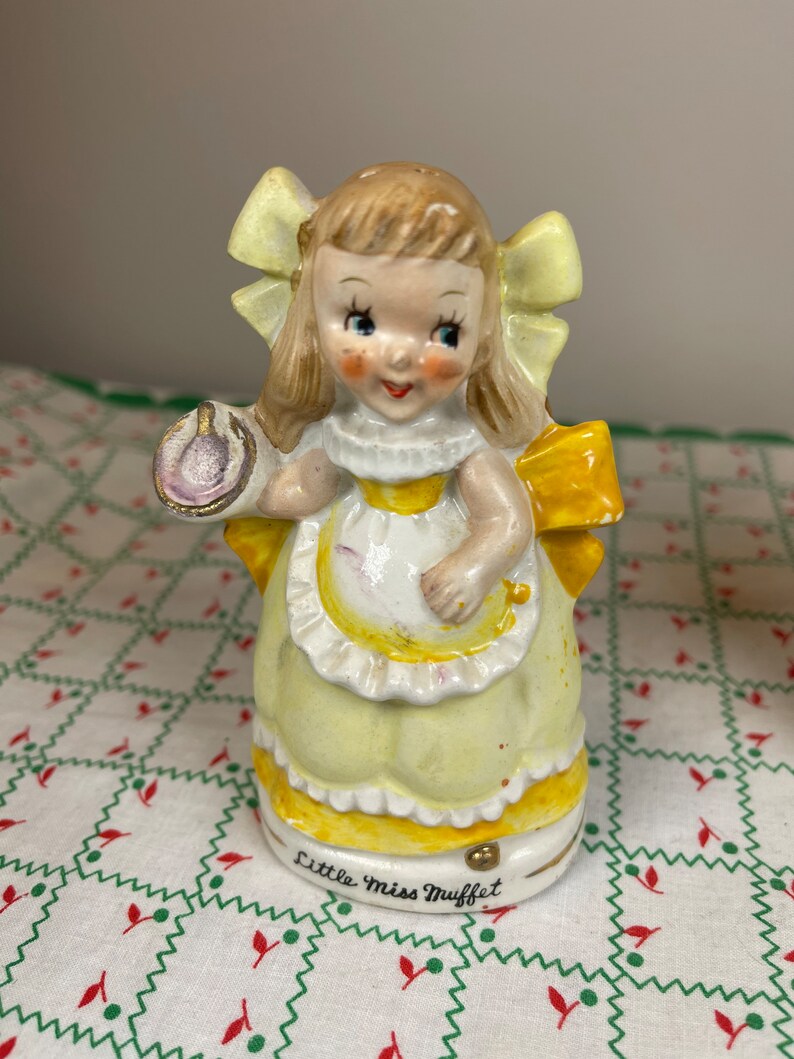 K&ouml;nnte beinhalten: Eine Porzellanfigur von Little Miss Muffet, einem M&auml;dchen in einem gelben Kleid und einer Sch&uuml;rze, das eine Sch&uuml;ssel und einen L&ouml;ffel h&auml;lt. Die Figur steht auf einem wei&szlig;en Hintergrund mit einem roten und gr&uuml;nen Muster.