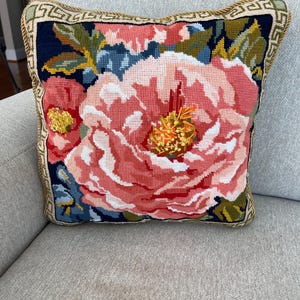 May include: Square decorative pillow with a large floral design. The pillow features a detailed needlepoint design with a large pink peony as the focal point, surrounded by blue and green leaves against a dark blue background. The edges are trimmed with a gold border.