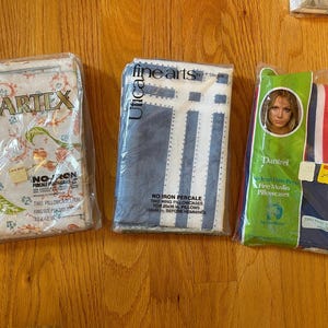 May include: Three packaged pillowcases on a wooden surface. One package is labeled "Martex" with a floral design. Another is "Utica" with blue and white stripes. The third is "Danirel" with red and blue stripes.