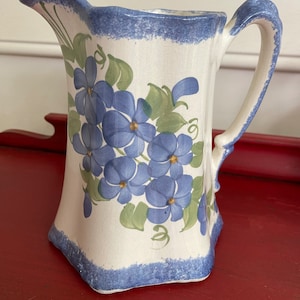 May include: A white ceramic pitcher with a blue rim and handle. The pitcher is decorated with a floral pattern of blue flowers and green leaves.