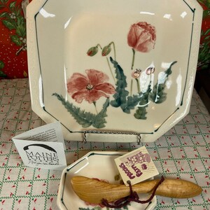 May include: A white ceramic octagonal serving dish with a green and brown floral design featuring red poppies. The dish is on a stand and next to it is a smaller white ceramic octagonal dish with a wooden butter knife. The smaller dish has a floral design and the text "Farm Fresh" on a tag.