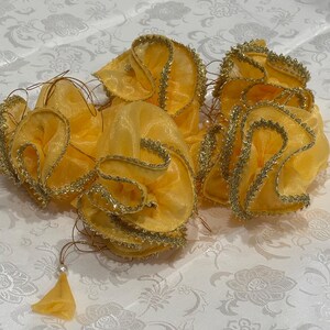 May include: A collection of yellow fabric flowers with gold trim and delicate string accents. Each flower is gathered and shaped, with a small tassel and pearl bead detail. The flowers are arranged on a white floral patterned surface.