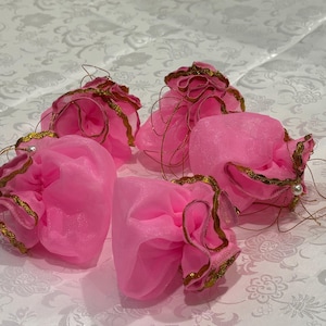 May include: Four pink organza gift bags with gold trim and delicate gold string accents. Each bag is gathered and has a ruffled edge. Small white pearls are attached to some of the bags. The bags are arranged on a white floral patterned surface.