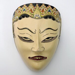 May include: A cream-colored, carved wooden mask with detailed facial features. The mask has dark brown eyes, a red mouth, and a decorative headdress with gold, black, and colorful geometric patterns. The mask is likely used for cultural performances.