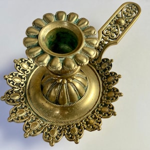 May include: Antique brass candle holder with a floral design. The candle holder features a round base with an ornate edge, a central cup for the candle, and a decorative handle. The brass has a slightly aged patina.