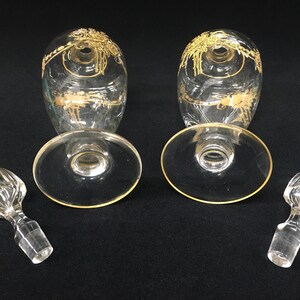 May include: Two clear glass wine glasses with gold floral designs and gold rims, along with two matching stoppers. The glasses are oval-shaped with a wide base. The stoppers are clear with a ribbed design.