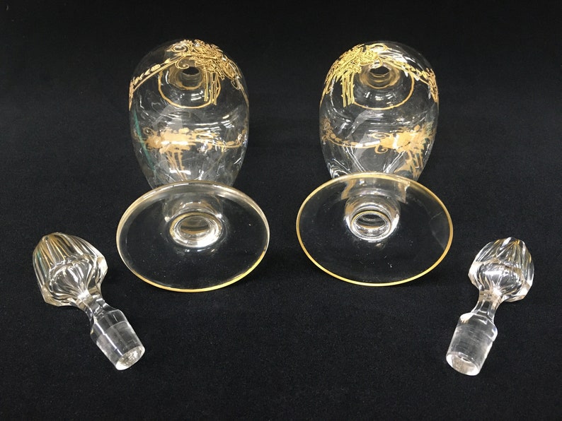 May include: Two clear glass wine glasses with gold floral designs and gold rims, along with two matching stoppers. The glasses are oval-shaped with a wide base. The stoppers are clear with a ribbed design.