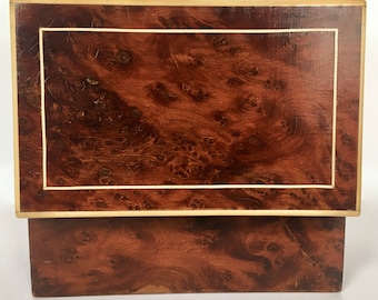 May include: A rectangular wooden box with a hinged lid. The box features a rich, dark brown burl wood grain pattern with a lighter wood border and gold trim. The box is likely used for storage or display.