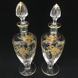 May include: Two clear glass perfume bottles with gold floral designs and gold trim. Each bottle has a clear stopper with gold accents. The bottles are displayed on a black surface.