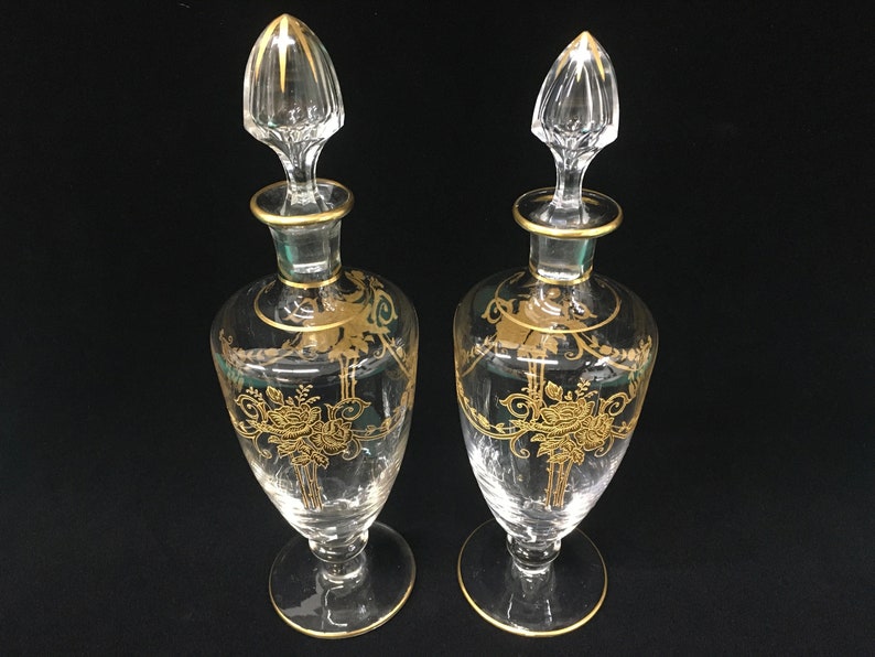 May include: Two clear glass perfume bottles with gold floral designs and gold trim. Each bottle has a clear stopper with gold accents. The bottles are displayed on a black surface.