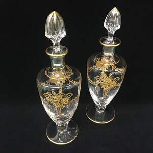 May include: Two clear glass perfume bottles with gold floral designs and gold trim. Each bottle has a clear stopper with gold accents. The bottles are on a black surface.