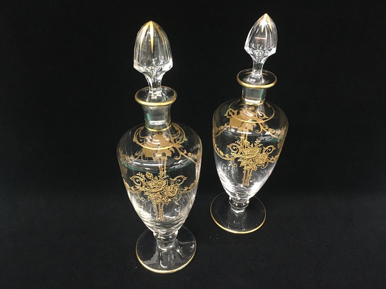 May include: Two clear glass perfume bottles with gold floral designs and gold trim. Each bottle has a clear stopper with gold accents. The bottles are on a black surface.