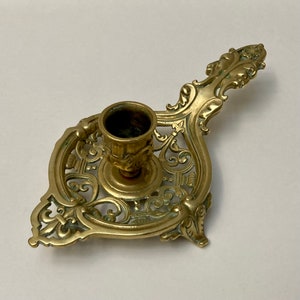 May include: Ornate brass candlestick holder with a handle. The candle holder features a detailed, openwork design with floral and scroll motifs. The cup for the candle is centered and has a decorative pattern. The brass has a slightly aged patina.