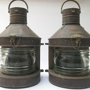 May include: Two antique copper and glass masthead lanterns. Each lantern has a clear glass globe, a copper top with a handle, and the word "MASTHEAD" is visible. The lanterns are mounted on a metal base.