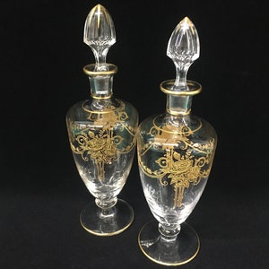 May include: Two clear glass perfume bottles with gold floral designs and gold trim. Each bottle has a clear stopper with a gold accent. The bottles are on a black background.