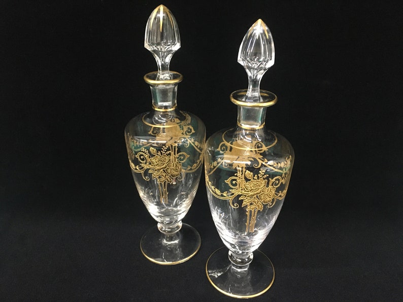 May include: Two clear glass perfume bottles with gold floral designs and gold trim. Each bottle has a clear stopper with a gold accent. The bottles are on a black background.
