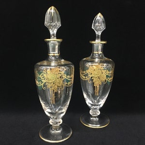 May include: Two clear glass perfume bottles with gold accents and stoppers. Each bottle features a gold floral design with green accents. The bottles have a round base and a long, slender body.
