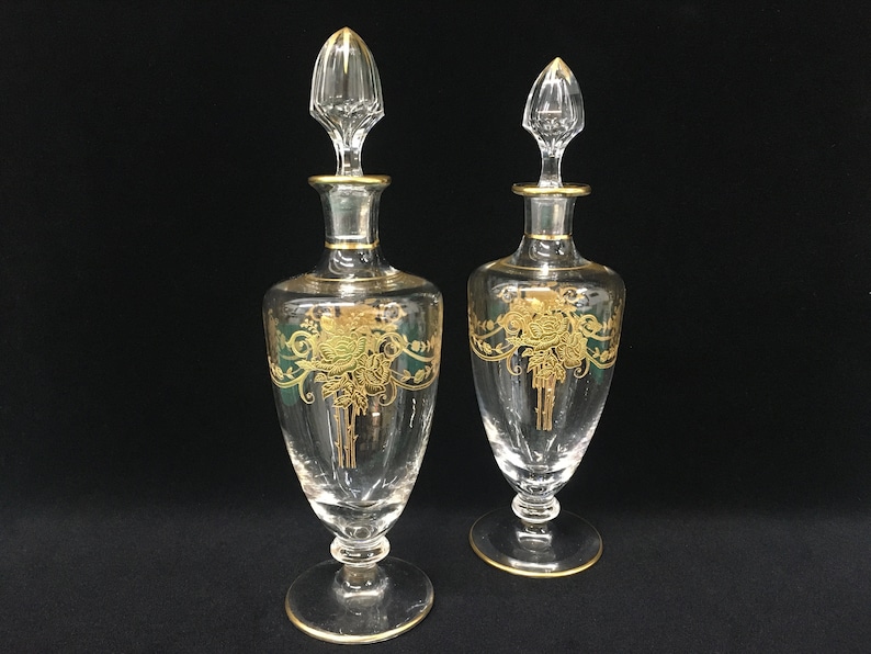 May include: Two clear glass perfume bottles with gold accents and stoppers. Each bottle features a gold floral design with green accents. The bottles have a round base and a long, slender body.