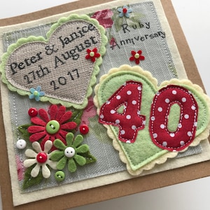 May include: A handmade Ruby Anniversary card with a floral design. The card features two heart-shaped appliqu&eacute;s with text and the number 40. Decorative felt flowers and buttons add detail. The card is on a neutral background.