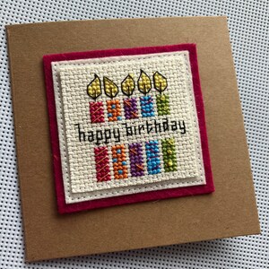 Peut inclure: Une carte faite &agrave; la main avec une bordure en feutre rouge et un fond blanc. La carte pr&eacute;sente un motif de point de croix de cinq bougies color&eacute;es avec des flammes et le texte "Joyeux anniversaire".