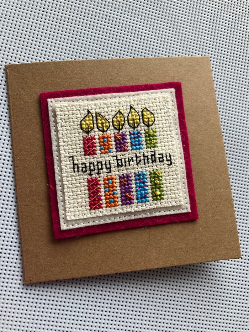 Peut inclure: Une carte faite &agrave; la main avec une bordure en feutre rouge et un fond blanc. La carte pr&eacute;sente un motif de point de croix de cinq bougies color&eacute;es avec des flammes et le texte "Joyeux anniversaire".
