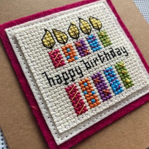 Peut inclure: Une carte faite &agrave; la main avec un motif de point de croix de bougies color&eacute;es et le texte "Happy Birthday". La carte est sur un fond brun avec une bordure rose.