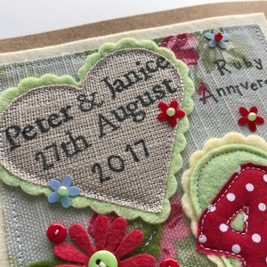 May include: Handmade Ruby Anniversary card with a heart-shaped burlap element. The card features the names "Peter & Janice" and the date "27th August 2017". Decorated with felt flowers and the words "Ruby Anniversary".