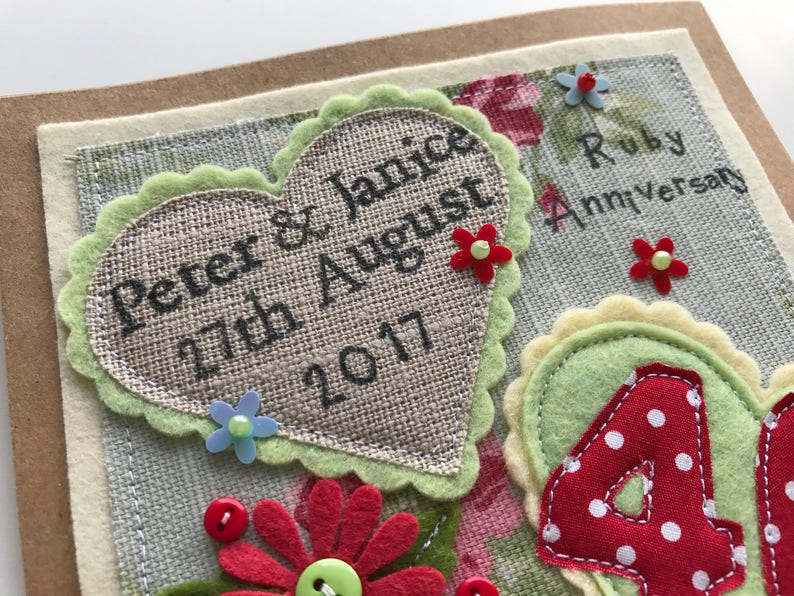 May include: Handmade Ruby Anniversary card with a heart-shaped burlap element. The card features the names "Peter & Janice" and the date "27th August 2017". Decorated with felt flowers and the words "Ruby Anniversary".