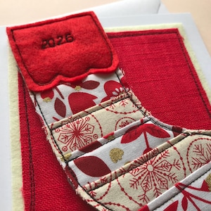 May include: A greetings card featuring a fabric appliqué of a Christmas stocking with the year 2026 embroidered on the red felt cuff. The stocking body is made of cream fabric with red and gold snowflake and floral patterns.
