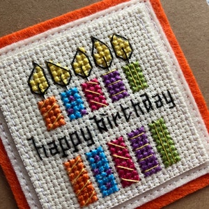 Peut inclure: Une carte blanche avec un motif de bougies color&eacute;es en point de croix et le texte "Happy Birthday". La carte est entour&eacute;e d'une double bordure en feutre orange.