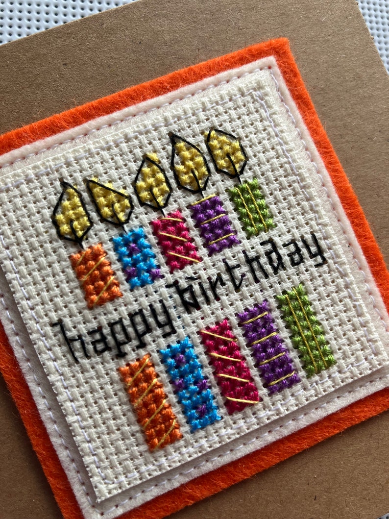 Peut inclure: Une carte blanche avec un motif de bougies color&eacute;es en point de croix et le texte "Happy Birthday". La carte est entour&eacute;e d'une double bordure en feutre orange.
