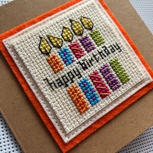 Peut inclure: Une carte faite &agrave; la main avec un motif de bougies color&eacute;es brod&eacute; au point de croix et le texte "Happy birthday". La carte est d&eacute;cor&eacute;e d'un fond en papier brun et d'une bordure en feutre orange.