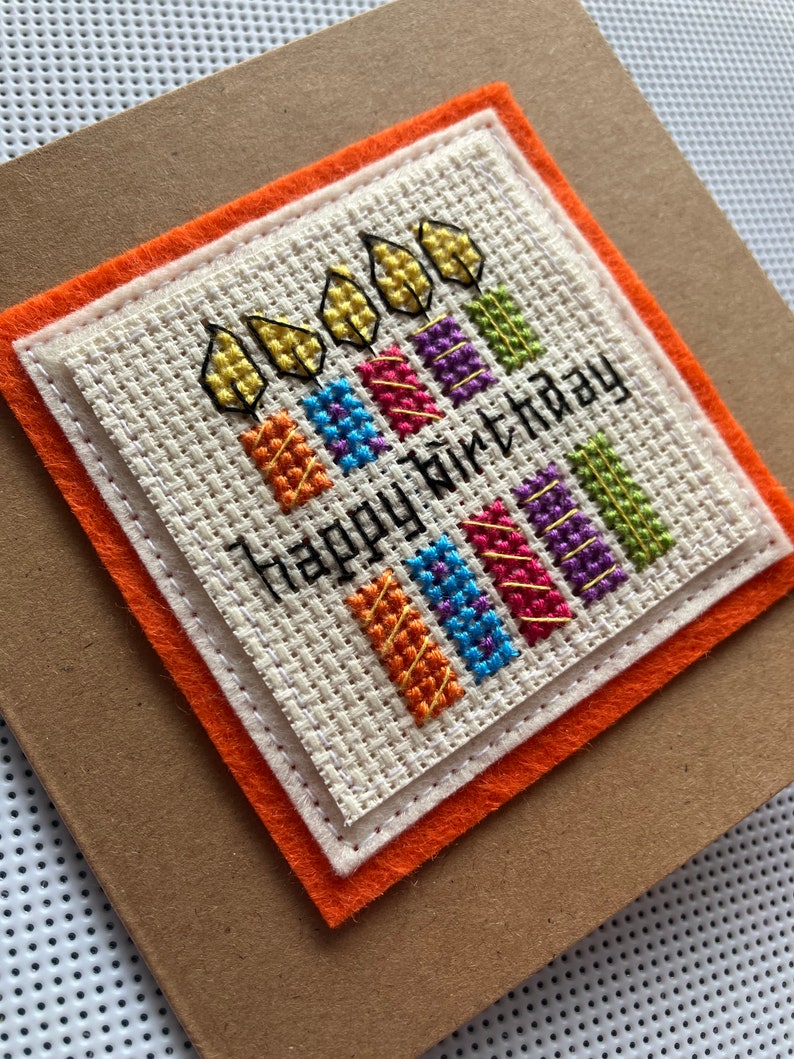 Peut inclure: Une carte faite &agrave; la main avec un motif de bougies color&eacute;es brod&eacute; au point de croix et le texte "Happy birthday". La carte est d&eacute;cor&eacute;e d'un fond en papier brun et d'une bordure en feutre orange.