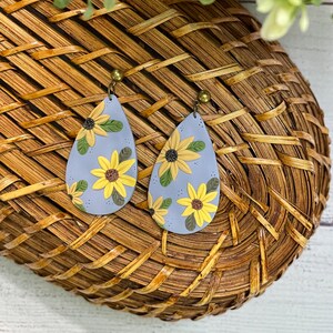 May include: A pair of teardrop-shaped earrings with a light blue background and a floral pattern of yellow sunflowers. The earrings are made of polymer clay and have gold-colored hooks.