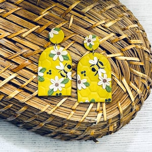 May include: A pair of yellow polymer clay earrings shaped like arched honeycombs with white flowers and bees. The earrings are hanging from a woven basket.