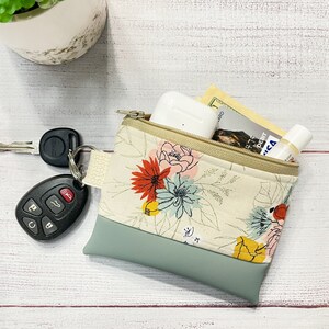 May include: A small zippered pouch with a floral print and a light blue bottom. The pouch is open and contains a pair of keys, a small white electronic device, a credit card, and a small tube of lotion.