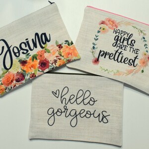 May include: Three beige canvas cosmetic bags with black zippers. One bag features the name "Josina" with floral accents. Another says "Happy girls are the prettiest" with a floral wreath. The third bag says "hello gorgeous".