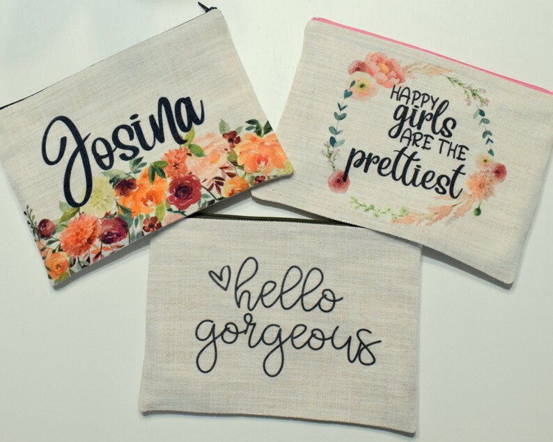May include: Three beige canvas cosmetic bags with black zippers. One bag features the name "Josina" with floral accents. Another says "Happy girls are the prettiest" with a floral wreath. The third bag says "hello gorgeous".