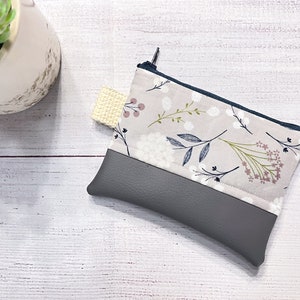 May include: A small gray and beige zippered pouch with a floral print. The pouch has a gray faux leather bottom and a beige fabric top with a white floral pattern.
