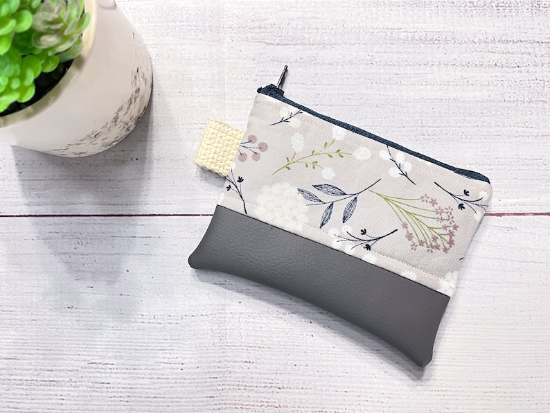 May include: A small gray and beige zippered pouch with a floral print. The pouch has a gray faux leather bottom and a beige fabric top with a white floral pattern.