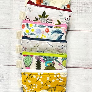 May include: A stack of six small zippered pouches with various floral and cactus prints. The pouches are made of fabric and have a zipper closure. The pouches are stacked on a white wooden surface.