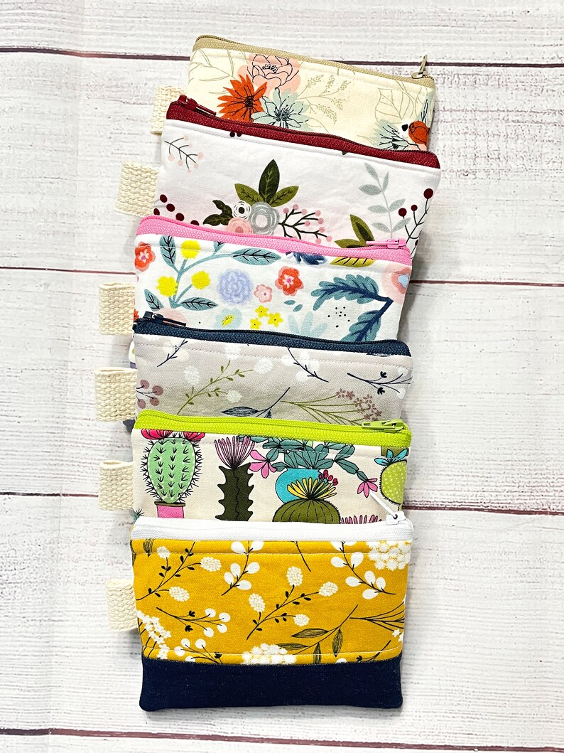 May include: A stack of six small zippered pouches with various floral and cactus prints. The pouches are made of fabric and have a zipper closure. The pouches are stacked on a white wooden surface.