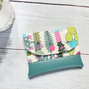 May include: A small, teal-colored zippered pouch with a flap closure. The flap is made of white fabric with a colorful cactus print. The pouch is perfect for storing small items like cash, cards, or jewelry.