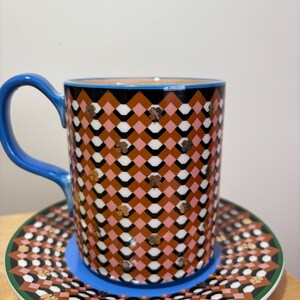 May include: A patterned ceramic coffee mug and saucer set. The mug has a blue handle and a geometric design in brown, pink, black, and white. The saucer mirrors the design with a green border and blue base.