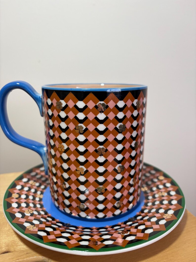 May include: A patterned ceramic coffee mug and saucer set. The mug has a blue handle and a geometric design in brown, pink, black, and white. The saucer mirrors the design with a green border and blue base.