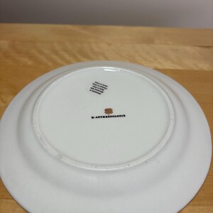 May include: White ceramic plate with a slightly raised rim. The underside features the text "by ANTHROPOLOGIE" in black, along with a small floral design and a sticker. The plate is on a light brown wooden surface.